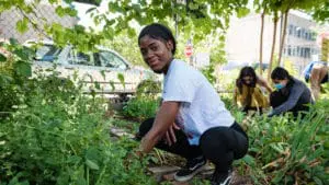 A young Black woman smiles towards the camera as she is squatting on the ground, surrounded by growing plants, at the Abib Newborn Learning Garden in New York City.