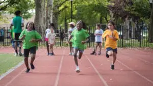Three smiling young kids of color run together on a track at a park in New York City at City Parks Foundation's free youth track and field program.