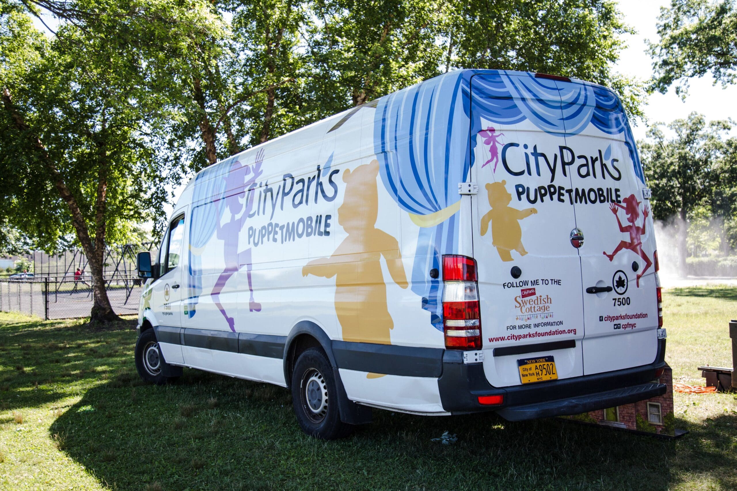 A 3/4 view of the CityParks PuppetMobile van. It was a large white van with illustrated graphics of puppets on the side and back. The CityParks PuppetMobile is adorned on the side and back. The van is parked in a sunny New York City park.