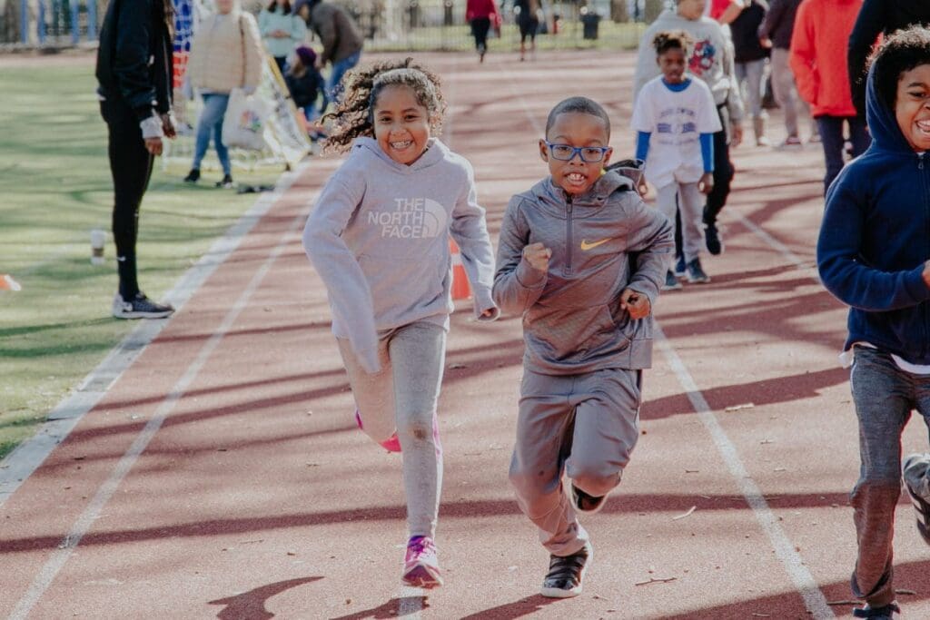 Two smiling young kids running at the City Parks Play free Jesse Owens Track and Field program in Harlem on a sunny day.