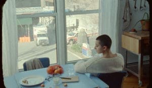 A still from the short film, "My Heart is My Only Country." It shows a person sitting at a table towards a window, with their head turned to the side.
