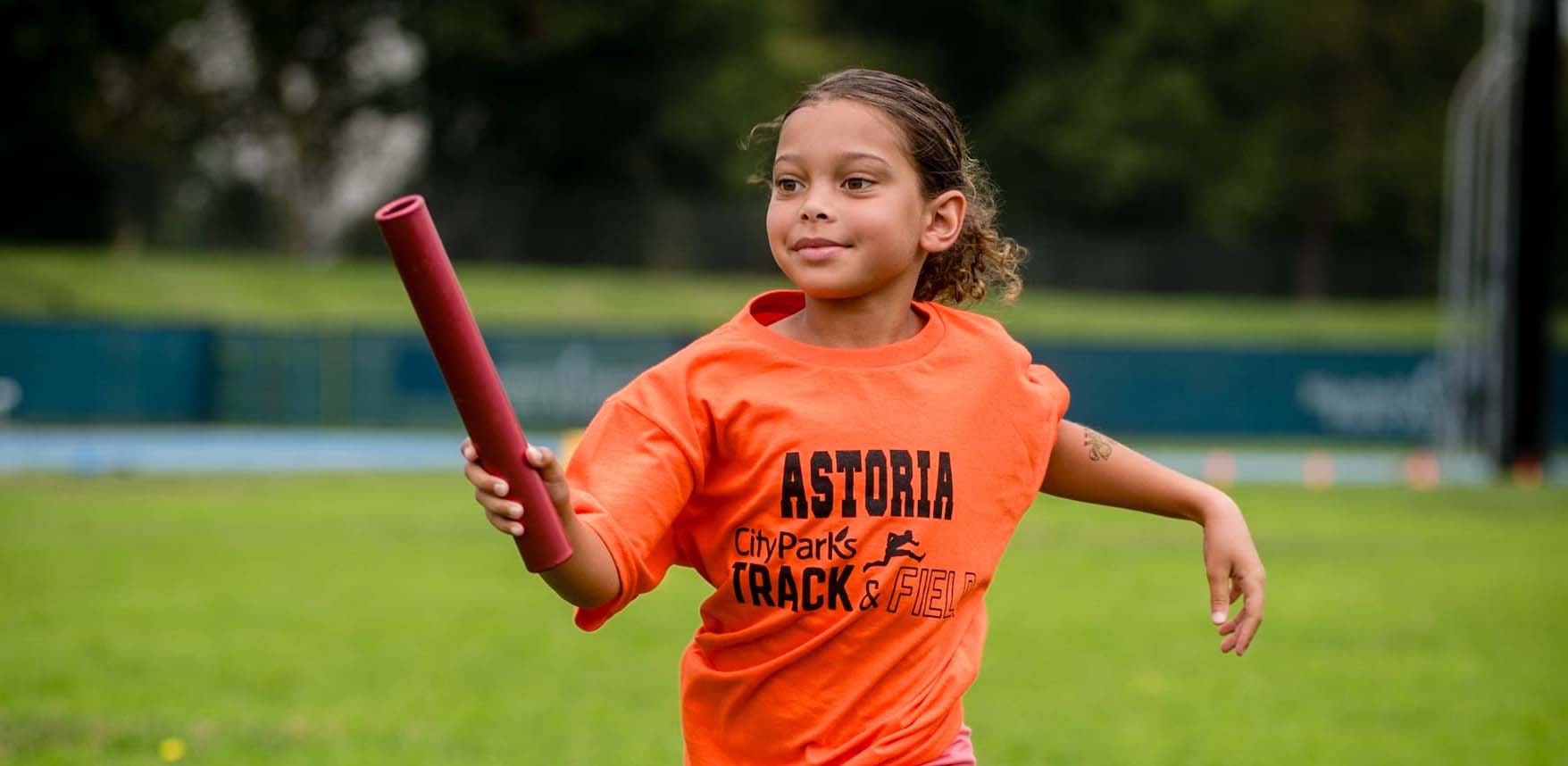 girl in orange shirt running with baton in a park