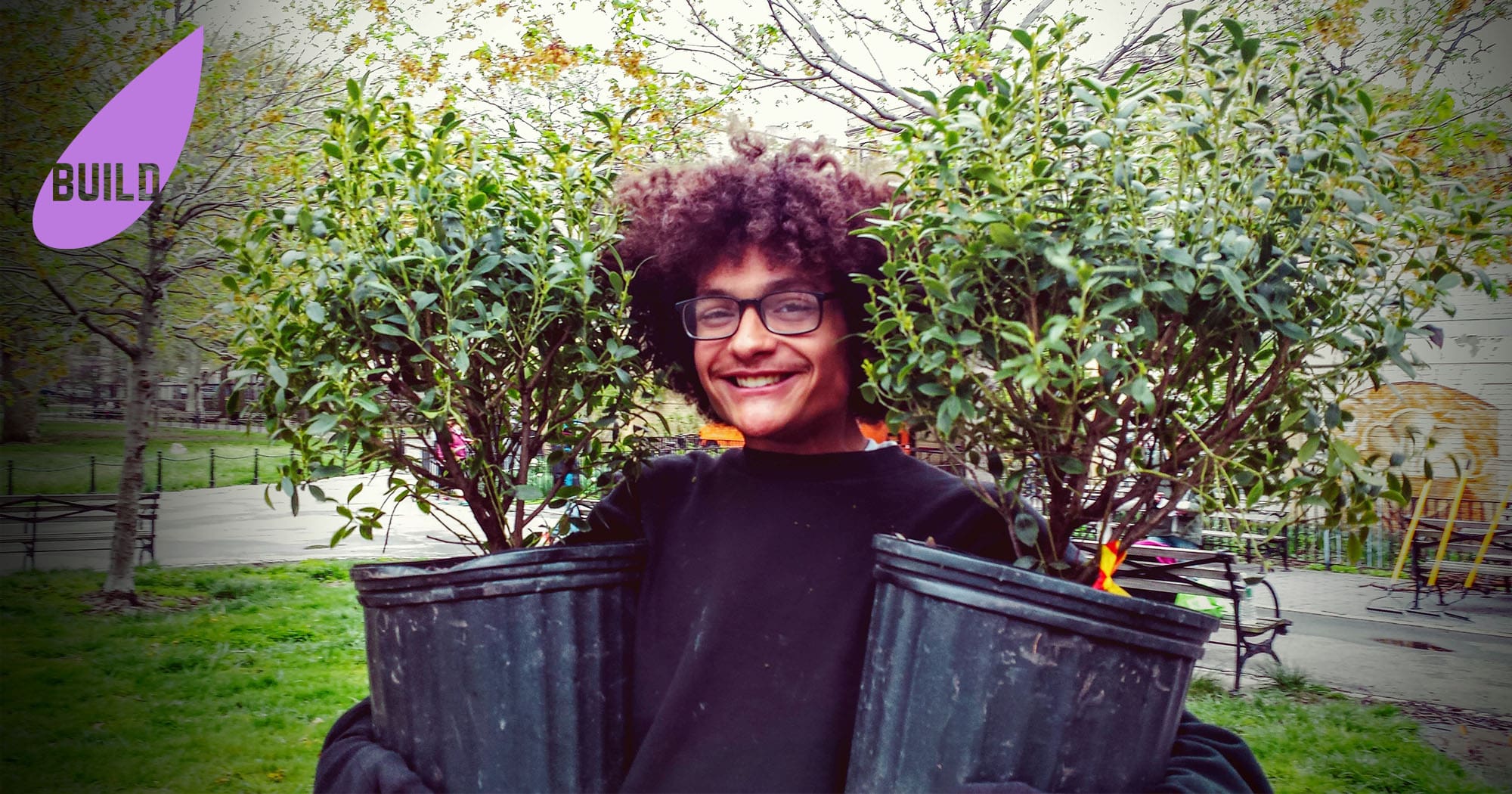 A smiling young Latinx volunteer holds two plants he is going to plant in a local New York City park. In the upper left hand corner is a purple leaf graphic with the word "BUILD" inside it.