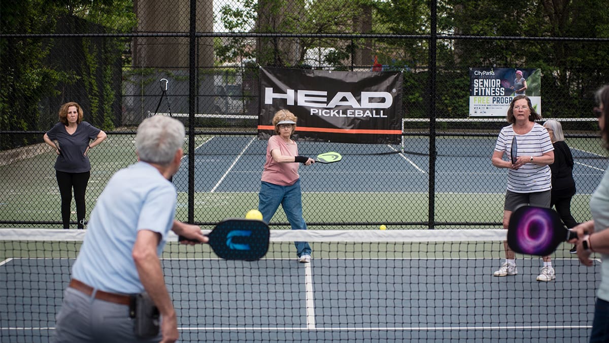 A group of four adults are playing pickleball on a tennis court. There are two women standing in the background.