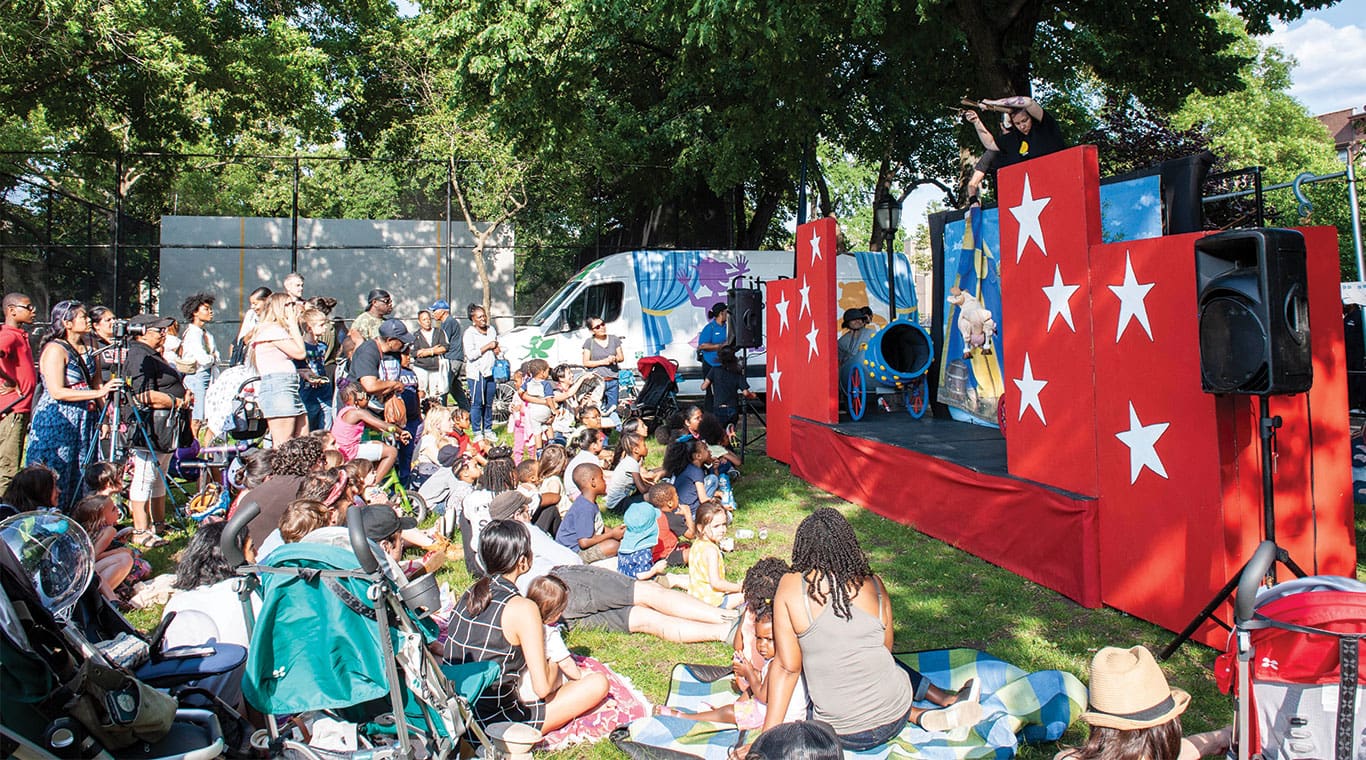 An audience of children and parents watch a free CityParks PuppetMobile performance in their local park. It is a bright summer day. Puppeteers are performing "Bessie's Big Shot!" to the crowd.