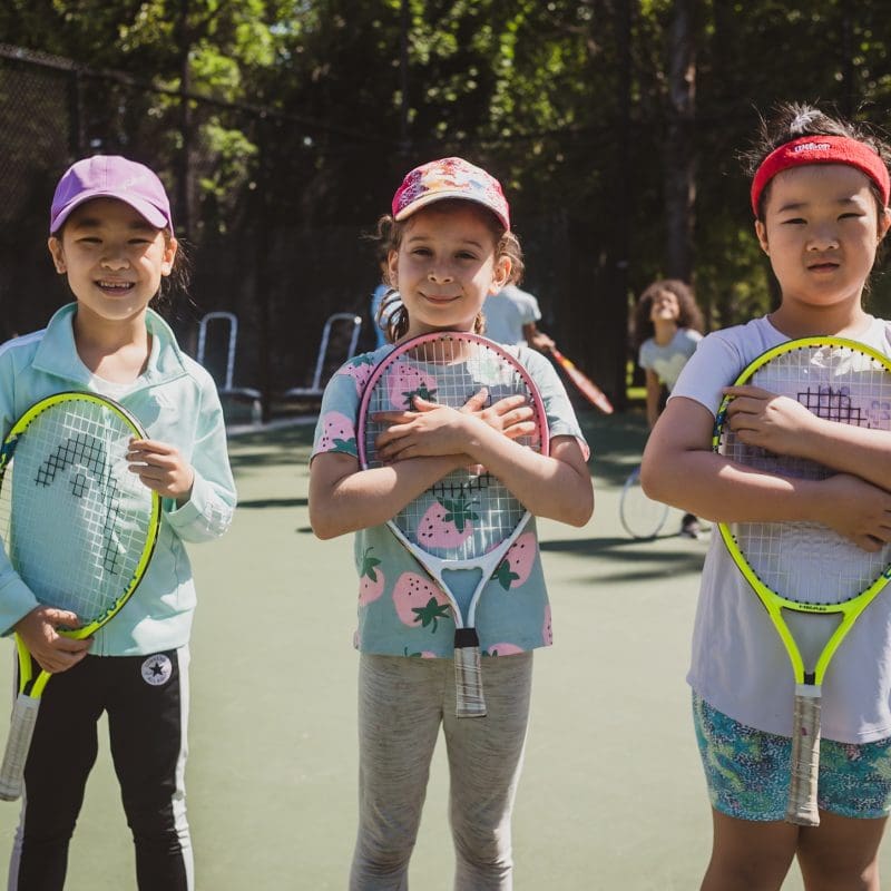 Three young girls wearing hats and holding tennis rackets in a park
