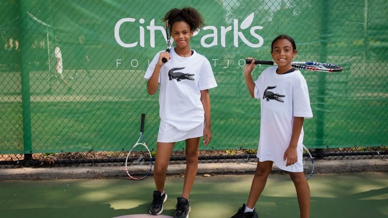 Two young black girls are standing with tennis racquets in their hands. They're facing the camera and smiling.