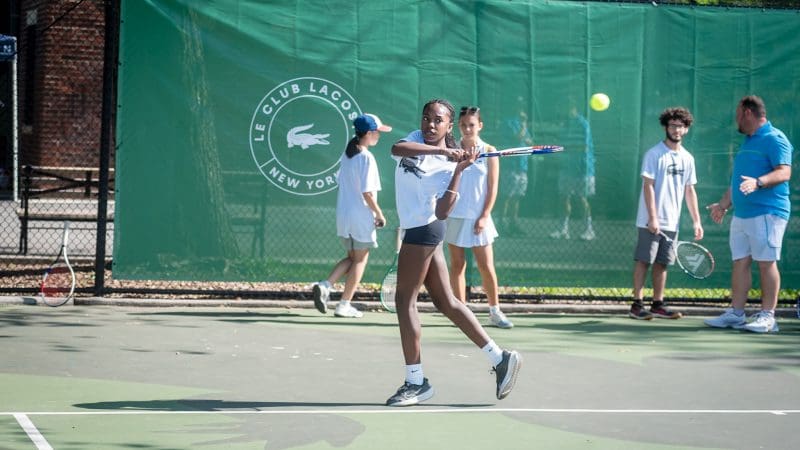 A young black girl is standing on a tennis court in the sun holding a tennis racquet mid-swing. Four people are standing behind her in the background.