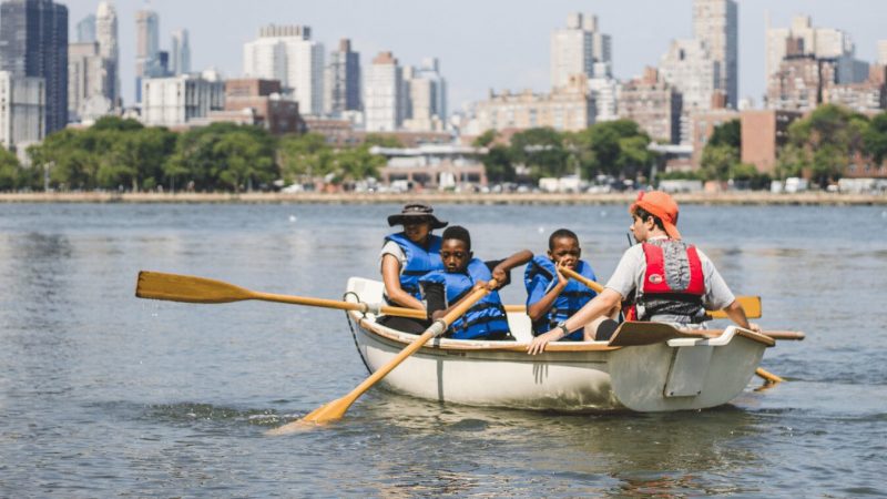 Three young boys and an adult are sitting in a row boat on a river in the city.