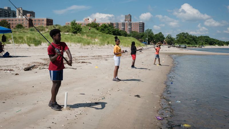 Four teenagers are standing on a beach and casting fishing lines into the water.