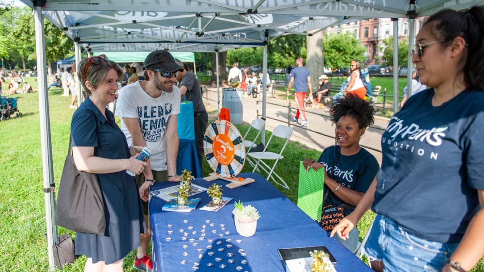 A smiling couple engages in conversation with two City Parks Foundation brand ambassadors at the info booth during a SummerStage show in Brooklyn.
