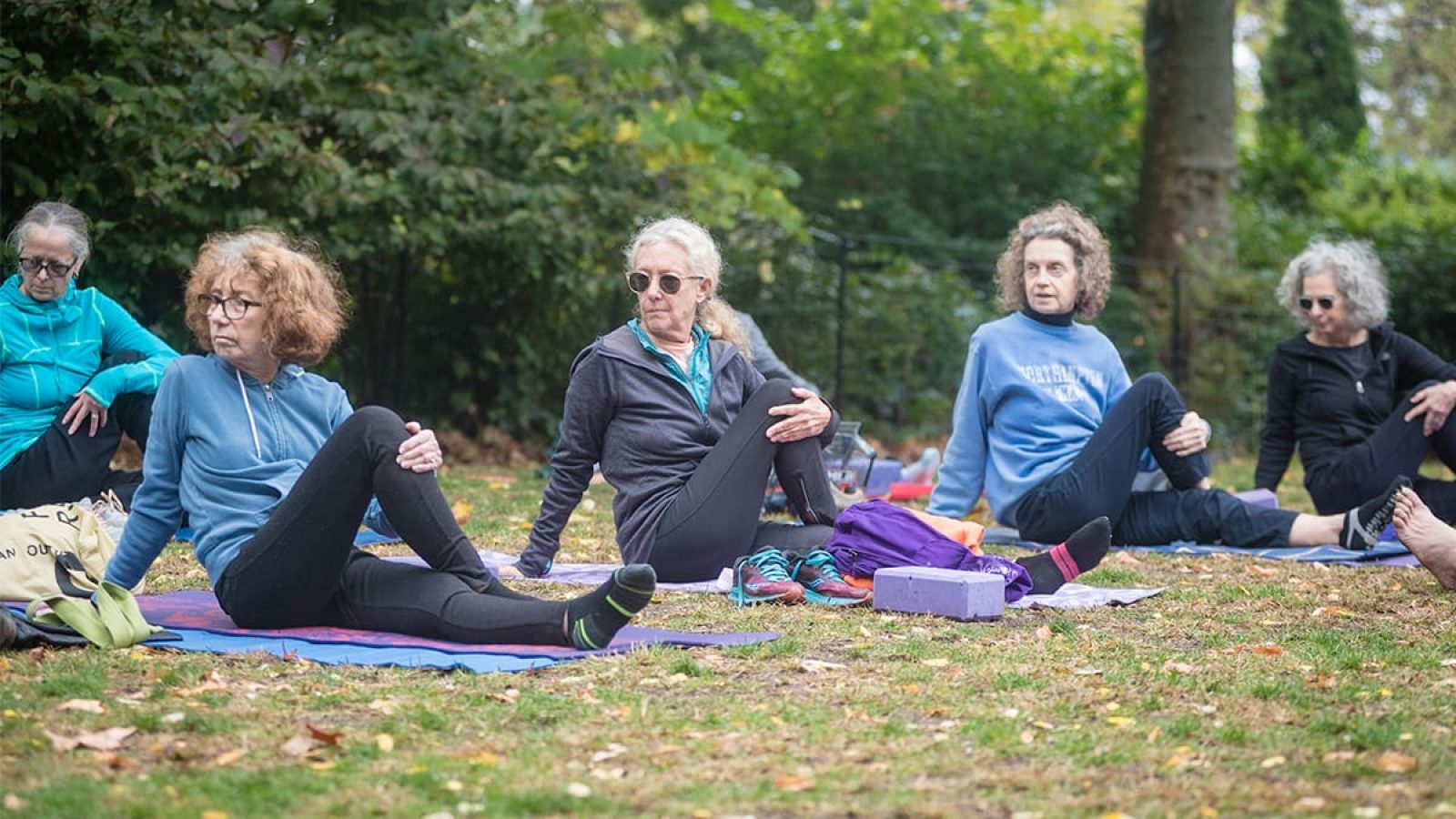 Four women are sitting in a row on a grassy lawn stretching one leg over their other leg.