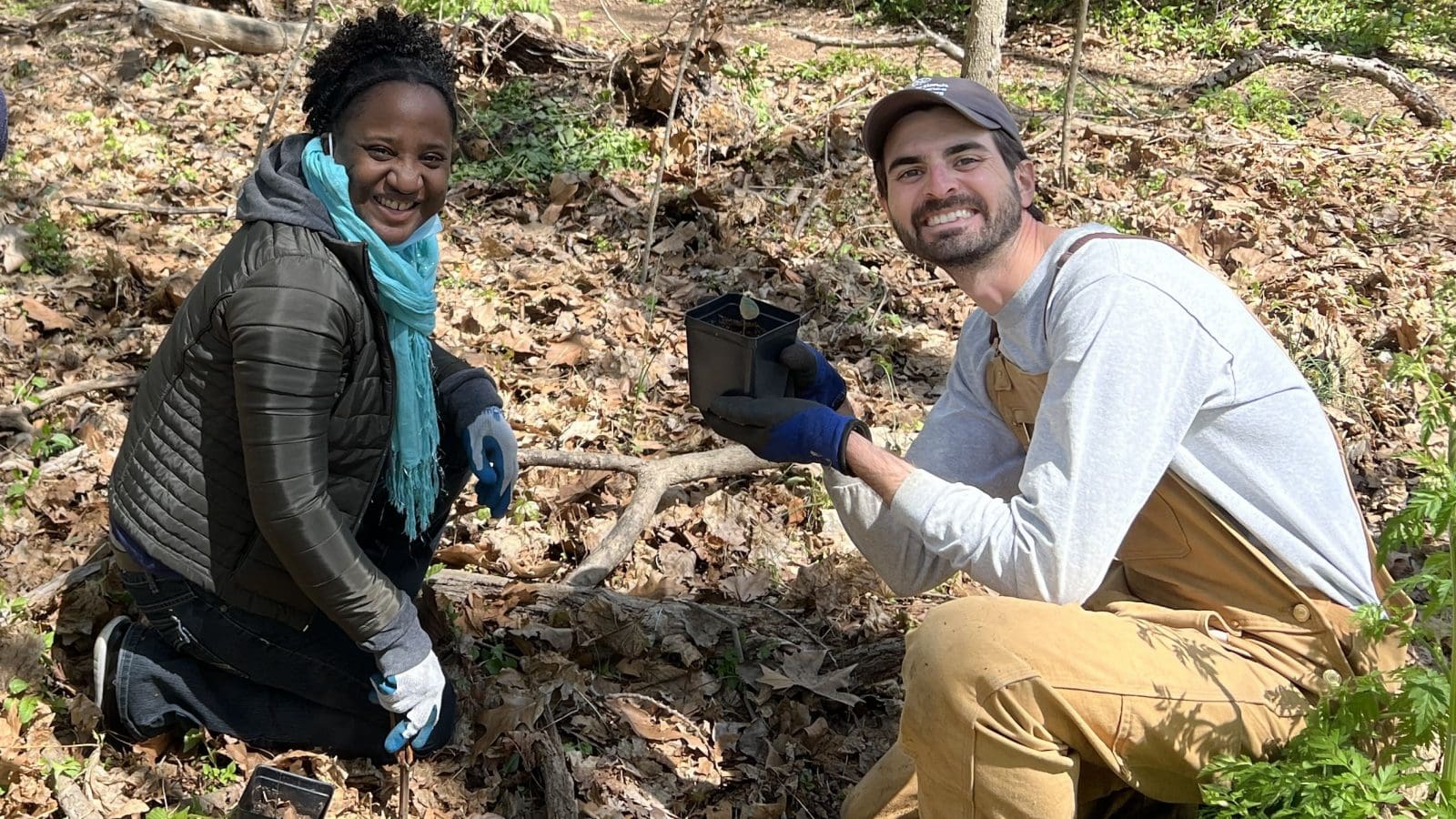 A black woman and a white man are kneeling on the ground and the man is holding a plant.