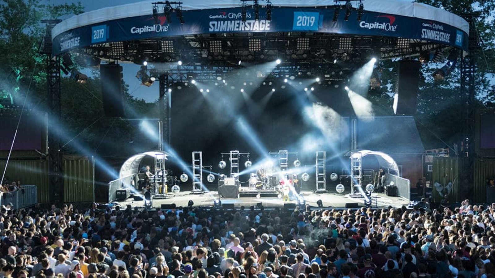 A stage wide shot of SummerStage in Central Park. There is a band on stage with various reflective lights shining across the stage and into the audience. There is a large crowd watching the show. It is evening, heading into sundown so there is a blue tone to the photo.