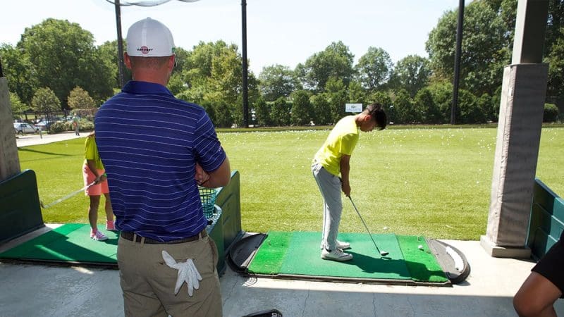 An adult man stands in the background while a young boy stands on a golf green getting ready to swing his golf club.