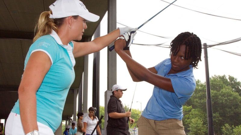 A woman is standing with a young boy. She is touching his wrist and talking to him about swinging his golf club.