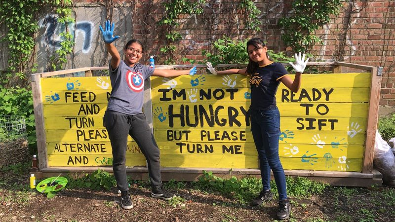 Two teenage girls are standing in a garden in front of a large composting bin that says "I'm not hungry, but please turn me."