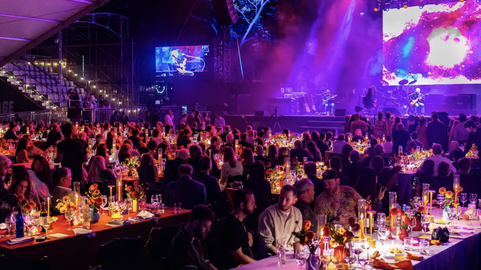 A large room full of people sitting at long tables with drinks and food. The room is illuminated with purple lighting.