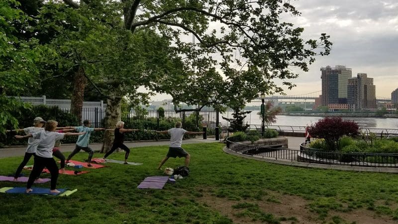 Five adults are doing yoga in a park on a grassy lawn. The city skyline and buildings are in the background