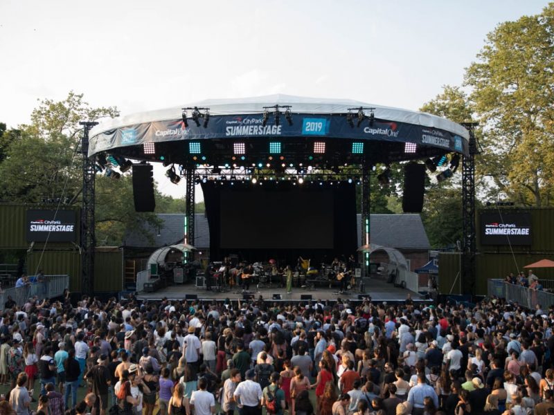 A stage wide shot of SummerStage in Central Park during the day. It is sunny and there is a large diverse crowd in front of the stage.
