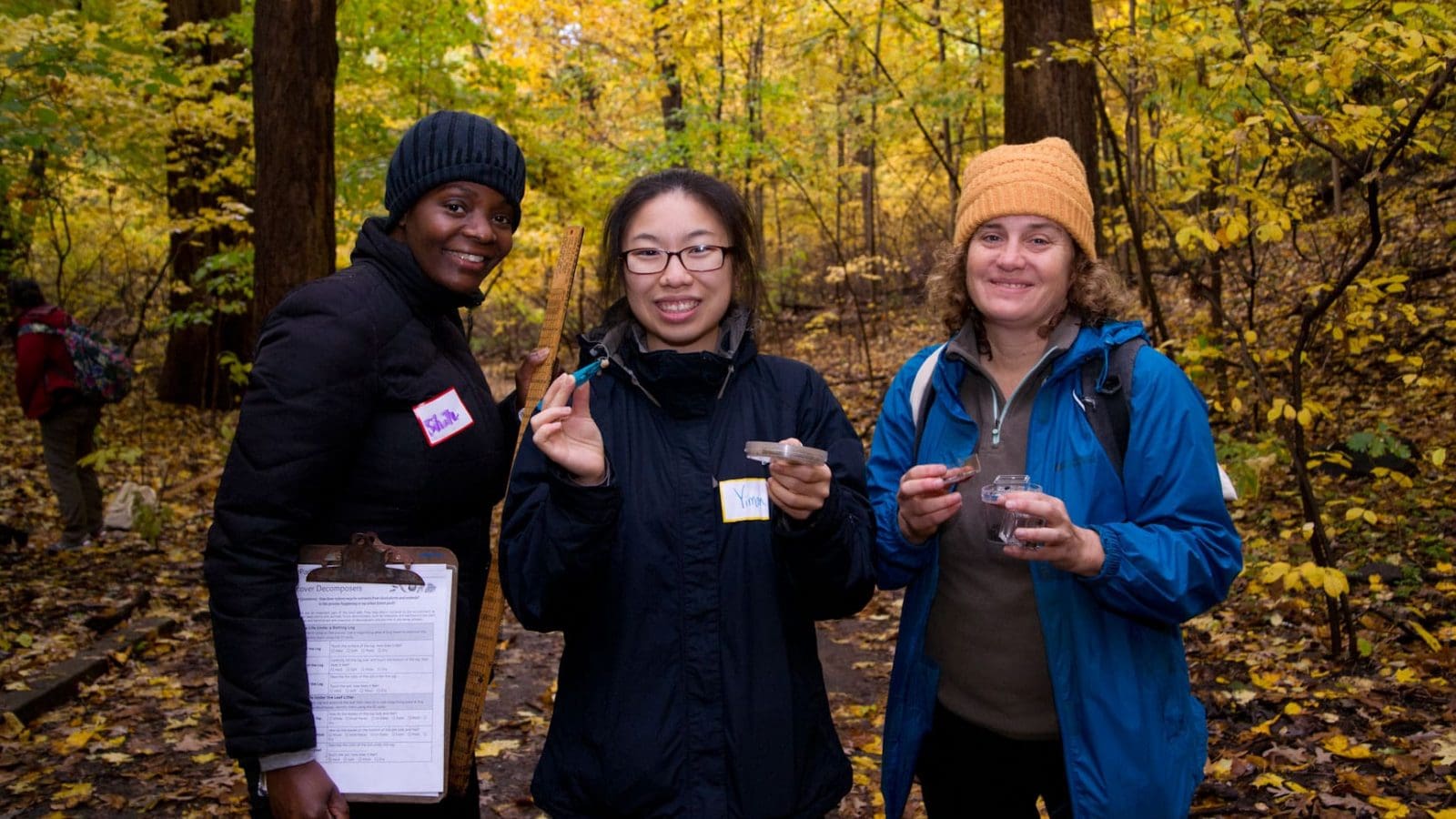 Three science teachers smiling in the woods holding clipboards and local insects through the outdoor Teacher Training program.