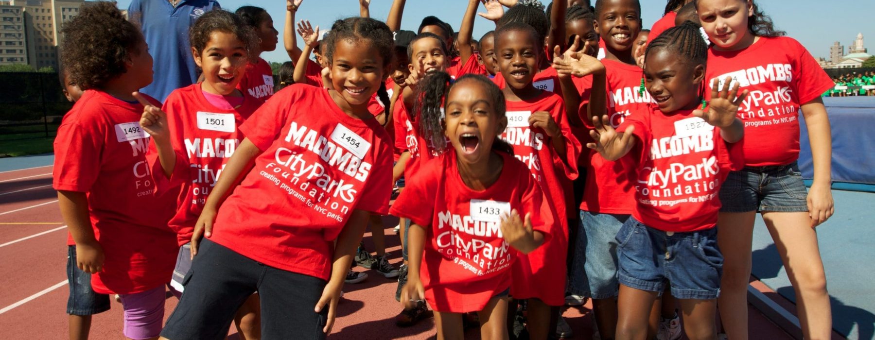 A group shot of elementary aged kids - mostly BIPOC - standing, smiling and posing together in a New York City park.