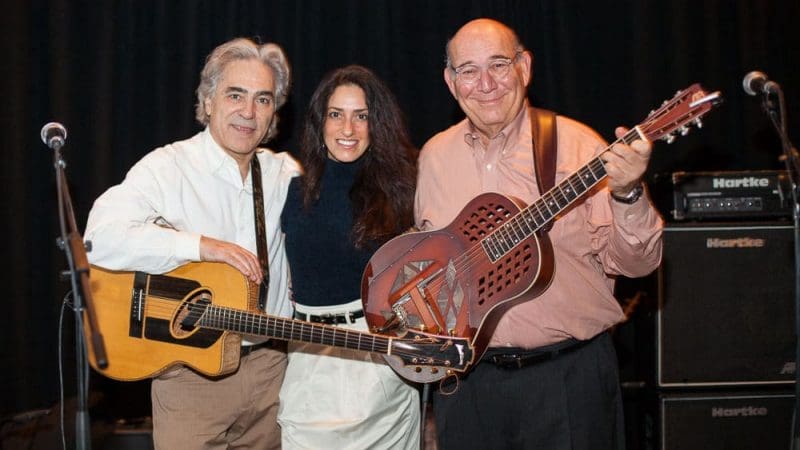 A woman is standing between two men holding guitars. The caption says "Woody Mann (left) with his wife Valerie Lettieri Mann and CPF Board Vice Chair, David Pinter"