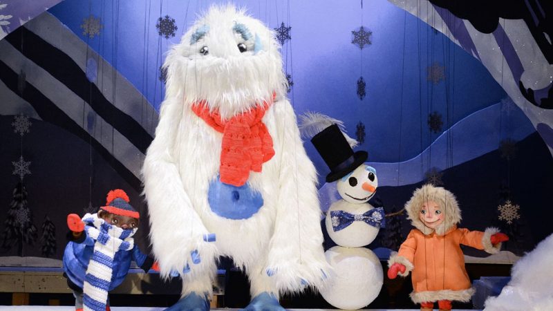 A photo from a performance of Yeti, Set, Snow! at the Swedish Cottage Marionette Theatre. It features a two young kids dressed in winter gear next to a snowman the tall fluffy white Yeti.