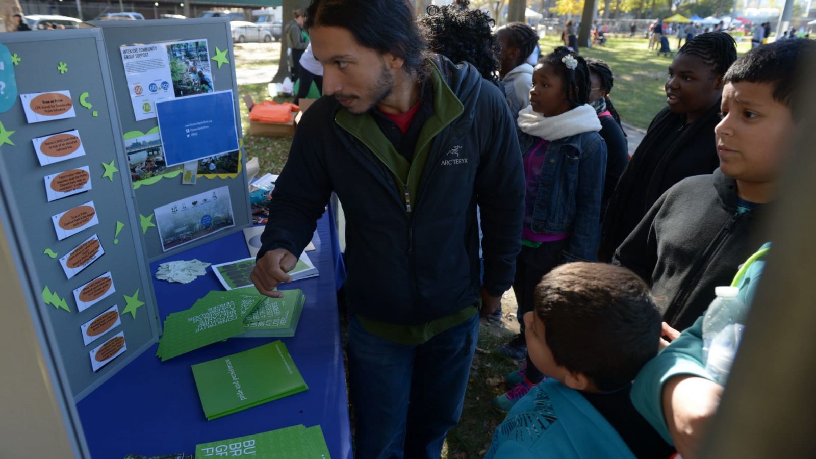 Community members engage with a Partnerships for Parks staff member at a community information booth. They are under a roofed-area in a New York City park.